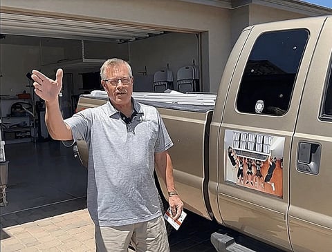 Man in gray shirt gesturing while standing next to a tan minivan in a driveway with garage in background